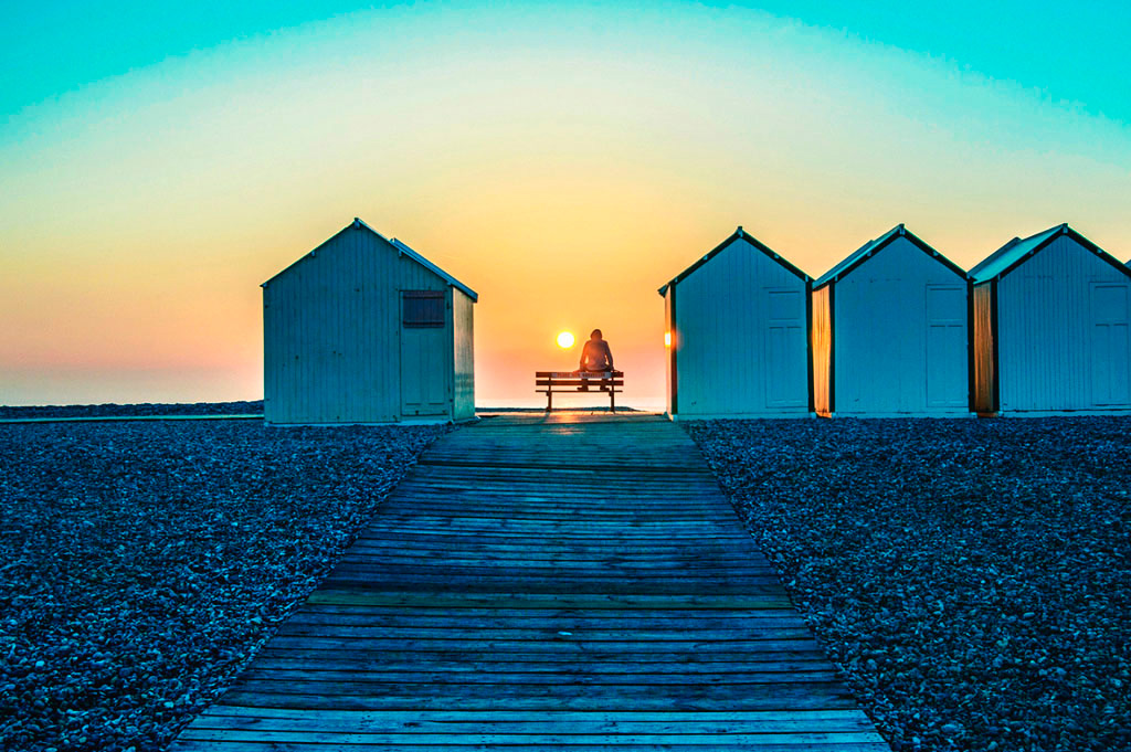 Photos Amiens, promeneur assis sur un banc entre les cabines de plage avec le soleil couchant qui iradie le paysage d'une couleur orange